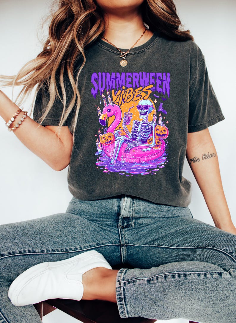 May include: Dark gray t-shirt with a graphic featuring a skeleton relaxing on a pink flamingo floatie, surrounded by pumpkins. The shirt has the text "Summerween Vibes" in purple and orange.
