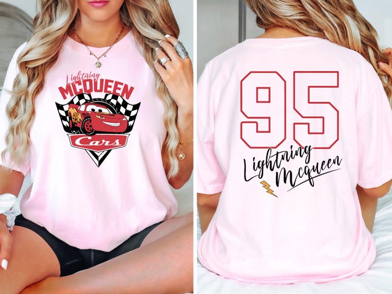May include: Pink t-shirt featuring a red cartoon racing car with the text "Lightning McQueen Cars" on the front. The back of the shirt displays the number "95" and the text "Lightning McQueen" in a cursive font.