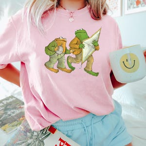 May include: A pink t-shirt with a graphic of two frogs. One frog is holding a kite and the other is holding a ball of yarn.