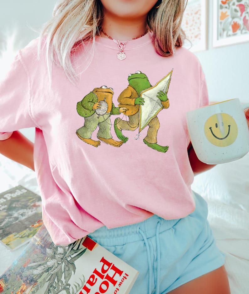 May include: A pink t-shirt with a graphic of two frogs. One frog is holding a kite and the other is holding a ball of yarn.