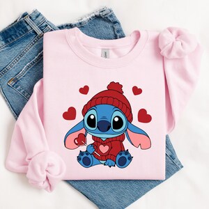 Pu&ograve; includere: Una felpa rosa chiaro con un personaggio dei cartoni animati, Stitch, che indossa un cappello e un maglione rossi, con in mano un lecca-lecca a forma di cuore. Cuori rossi sono sparsi intorno al personaggio. La felpa &egrave; appoggiata su un paio di jeans blu.