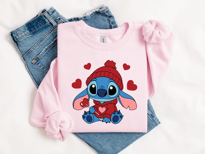 Pu&ograve; includere: Una felpa rosa chiaro con un personaggio dei cartoni animati, Stitch, che indossa un cappello e un maglione rossi, con in mano un lecca-lecca a forma di cuore. Cuori rossi sono sparsi intorno al personaggio. La felpa &egrave; appoggiata su un paio di jeans blu.