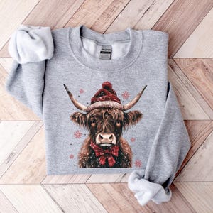 May include: A gray sweatshirt with a festive design featuring a Highland cow wearing a red Santa hat and a red bow tie. The cow is surrounded by snowflakes.