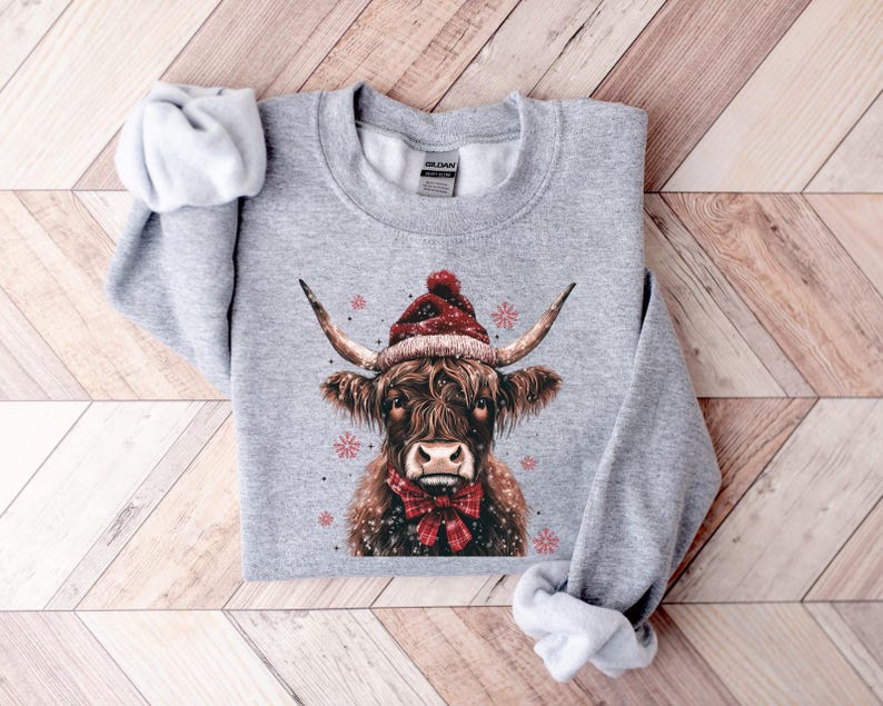 May include: A gray sweatshirt with a festive design featuring a Highland cow wearing a red Santa hat and a red bow tie. The cow is surrounded by snowflakes.