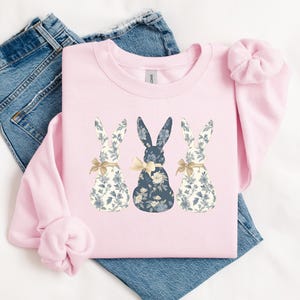 K&ouml;nnte beinhalten: Ein rosa Sweatshirt mit einem Design aus drei floral gemusterten Hasen. Die Hasen sind in einer Reihe angeordnet, wobei der mittlere Hase blau und die &auml;u&szlig;eren Hasen wei&szlig; sind.