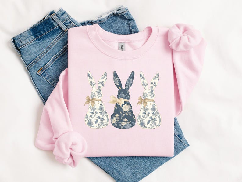 K&ouml;nnte beinhalten: Ein rosa Sweatshirt mit einem Design aus drei floral gemusterten Hasen. Die Hasen sind in einer Reihe angeordnet, wobei der mittlere Hase blau und die &auml;u&szlig;eren Hasen wei&szlig; sind.