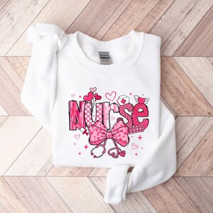 May include: White sweatshirt with a pink and white graphic design that says "Nurse" with hearts, a stethoscope, and a bandage.