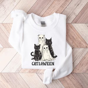 May include: White sweatshirt featuring a graphic of four cats, two in ghost costumes. The cats are black and white. The word "CATLOWEEN" is printed in black below the cats. The sweatshirt is made of a soft material.
