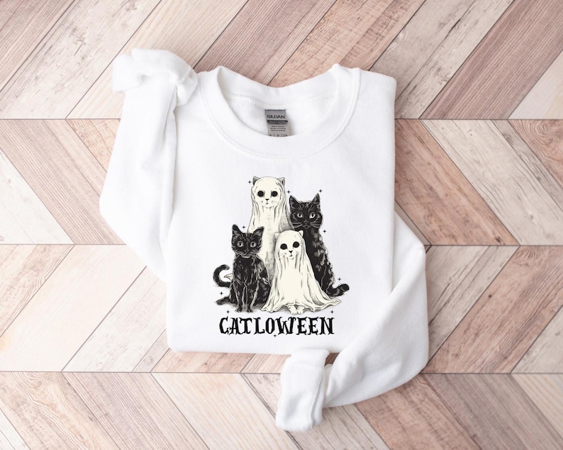 May include: White sweatshirt featuring a graphic of four cats, two in ghost costumes. The cats are black and white. The word "CATLOWEEN" is printed in black below the cats. The sweatshirt is made of a soft material.