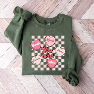 May include: Green sweatshirt with a checkered pattern and pink hearts that say "Loved", "Enough", "Strong", "Capable", and "Worthy". The text "You are" is in the center of the hearts.