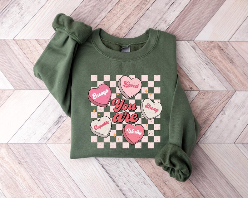 May include: Green sweatshirt with a checkered pattern and pink hearts that say "Loved", "Enough", "Strong", "Capable", and "Worthy". The text "You are" is in the center of the hearts.