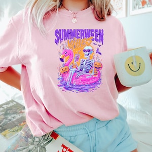 May include: Pink t-shirt with a graphic featuring a skeleton relaxing on a flamingo floatie, surrounded by pumpkins and bats. The text "SUMMERWEEN VIBES" is displayed above the graphic. A white mug with a yellow smiley face is held in the hand.
