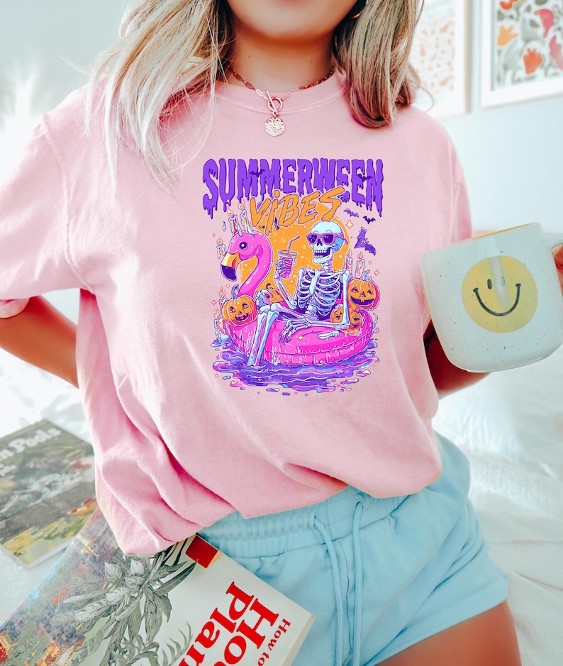 May include: Pink t-shirt with a graphic featuring a skeleton relaxing on a flamingo floatie, surrounded by pumpkins and bats. The text "SUMMERWEEN VIBES" is displayed above the graphic. A white mug with a yellow smiley face is held in the hand.
