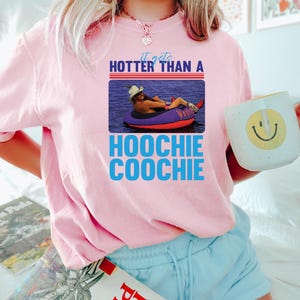 May include: Pink sweatshirt with the text "it gets HOTTER THAN A HOOCHIE COOCHIE" and a graphic of a person in a tube. The person is wearing a cowboy hat and is floating in the water. A white mug with a smiley face is held in the hand.