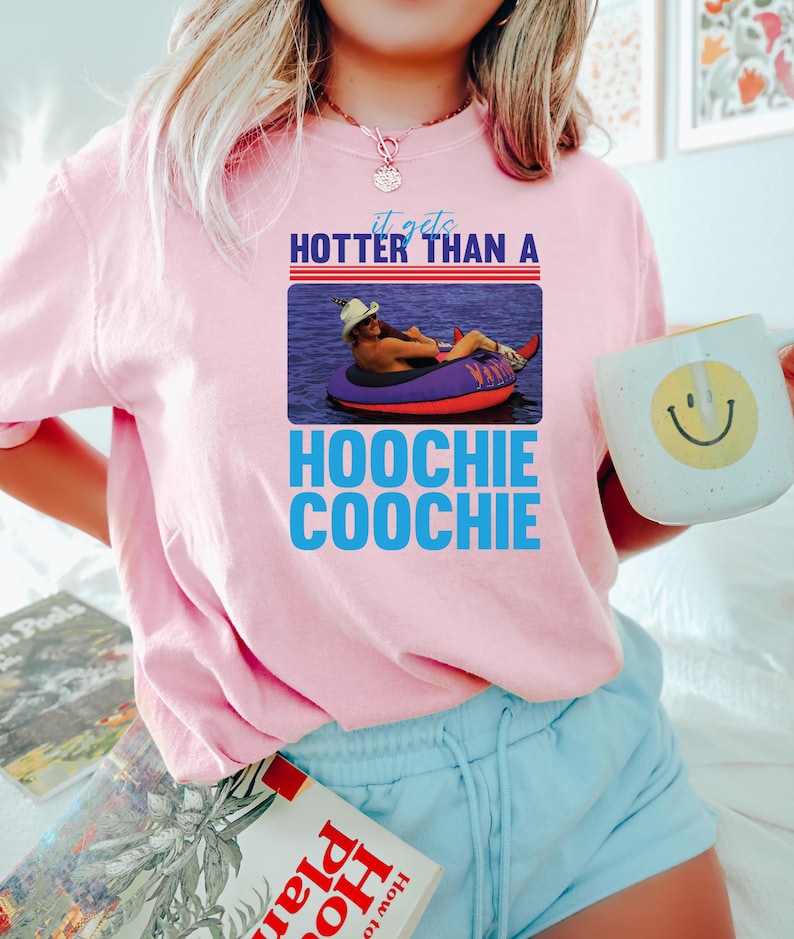 May include: Pink sweatshirt with the text "it gets HOTTER THAN A HOOCHIE COOCHIE" and a graphic of a person in a tube. The person is wearing a cowboy hat and is floating in the water. A white mug with a smiley face is held in the hand.