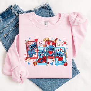 May include: Pink sweatshirt with a graphic print of Stitch from Lilo & Stitch. Stitch is shown in four different poses with hearts and the word "LOVE" on the sweatshirt.