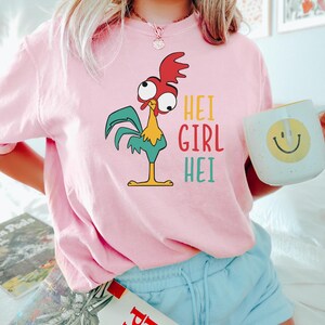 May include: Pink t-shirt with a cartoon rooster and the text "Hei Girl Hei" printed on it. The person wearing the shirt is holding a white mug with a yellow smiley face.
