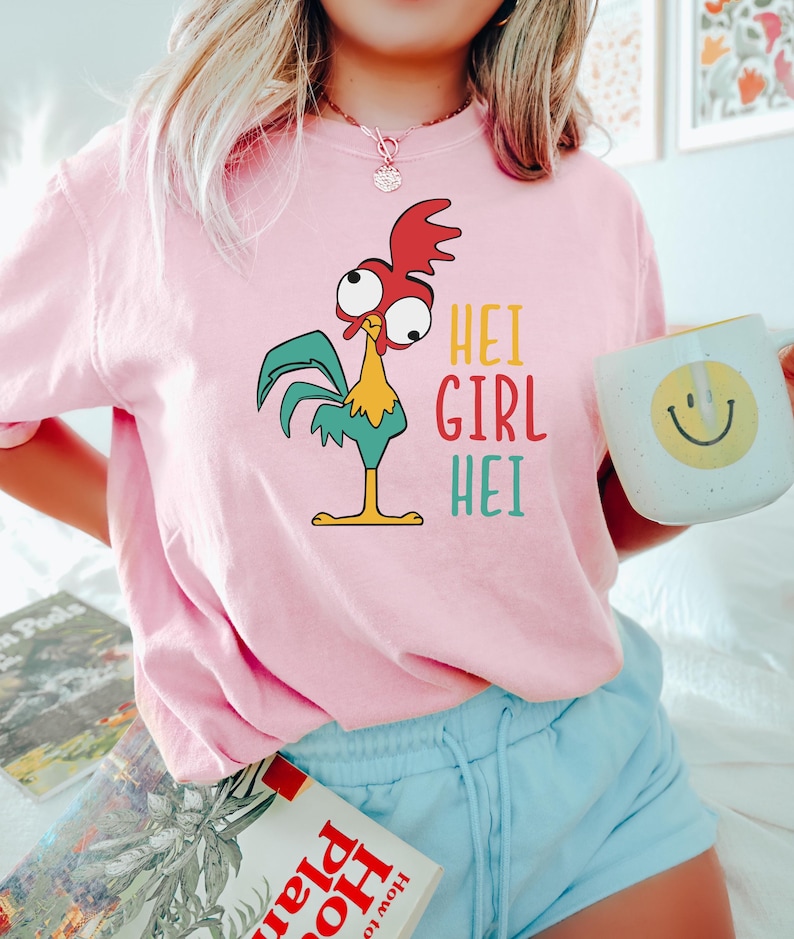 May include: Pink t-shirt with a cartoon rooster and the text "Hei Girl Hei" printed on it. The person wearing the shirt is holding a white mug with a yellow smiley face.