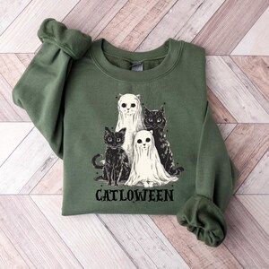 May include: Green sweatshirt featuring a graphic of four cats, two in black and two in white ghost costumes. The word "CATLOWEEN" is printed in black below the cats. The sweatshirt is made of a soft material.