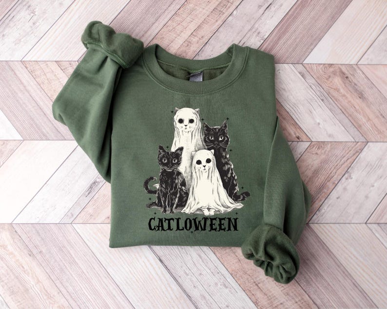 May include: Green sweatshirt featuring a graphic of four cats, two in black and two in white ghost costumes. The word "CATLOWEEN" is printed in black below the cats. The sweatshirt is made of a soft material.