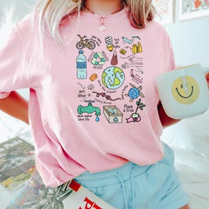 May include: Pink t-shirt with a graphic design of various ways to help the planet. The graphic includes a bicycle, a lightbulb, a water bottle, a faucet, a tree, a turtle, and the words "Recycle, Reduce, Reuse, Save the Planet, Plant a Tree, Don't Litter, Save Water, Save Life, Skip a Straw, Volunteer, Bike More, Drive Less, Turn Off Lights".