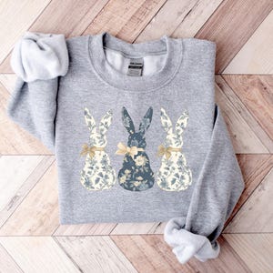 K&ouml;nnte beinhalten: Grauer Pullover mit drei floralen Hasen in einer Reihe. Die Hasen sind wei&szlig;, blau und wei&szlig; mit gelben Schleifen.