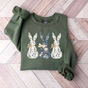 K&ouml;nnte beinhalten: Ein gr&uuml;nes Sweatshirt mit einem floralen Druck von drei Hasen. Die Hasen stehen in einer Reihe, wobei der mittlere Hase nach vorne schaut und die beiden anderen Hasen seitw&auml;rts schauen. Die Hasen tragen Schleifen.