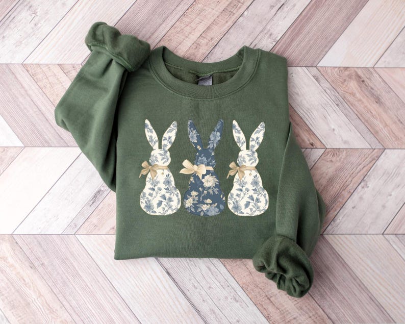 K&ouml;nnte beinhalten: Ein gr&uuml;nes Sweatshirt mit einem floralen Druck von drei Hasen. Die Hasen stehen in einer Reihe, wobei der mittlere Hase nach vorne schaut und die beiden anderen Hasen seitw&auml;rts schauen. Die Hasen tragen Schleifen.