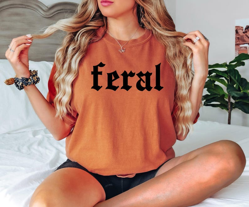 May include: A woman wearing a rust-coloured t-shirt with the word "feral" printed in black gothic lettering.