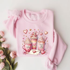 May include: Pink sweatshirt with a graphic of three mugs with whipped cream and hearts. The mugs say "XOXO", "Hugs and Kisses", and have hearts and other Valentine's Day themed designs.