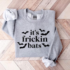 K&ouml;nnte beinhalten: Grauer Pullover mit dem Text "it's frickin' bats" in schwarzer Schrift und einer schwarzen Fledermausgrafik.