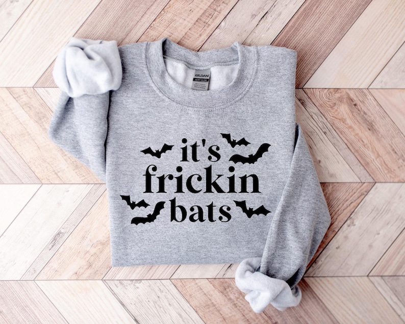 K&ouml;nnte beinhalten: Grauer Pullover mit dem Text "it's frickin' bats" in schwarzer Schrift und einer schwarzen Fledermausgrafik.