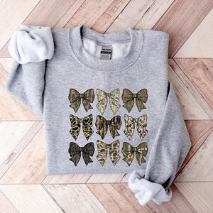 May include: A gray sweatshirt with a pattern of nine camouflage bows printed on the front. The bows are in shades of brown, green, and tan.