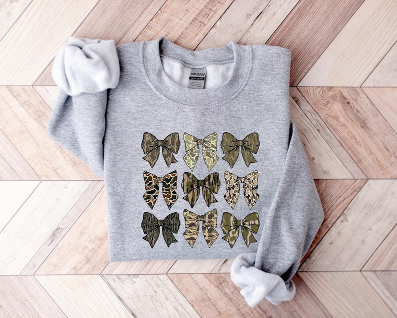 May include: A gray sweatshirt with a pattern of nine camouflage bows printed on the front. The bows are in shades of brown, green, and tan.