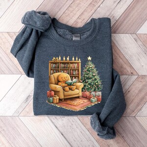 May include: A dark blue sweatshirt with a Christmas-themed design featuring a gingerbread person sitting in a chair reading a book by a fireplace with a Christmas tree and presents.
