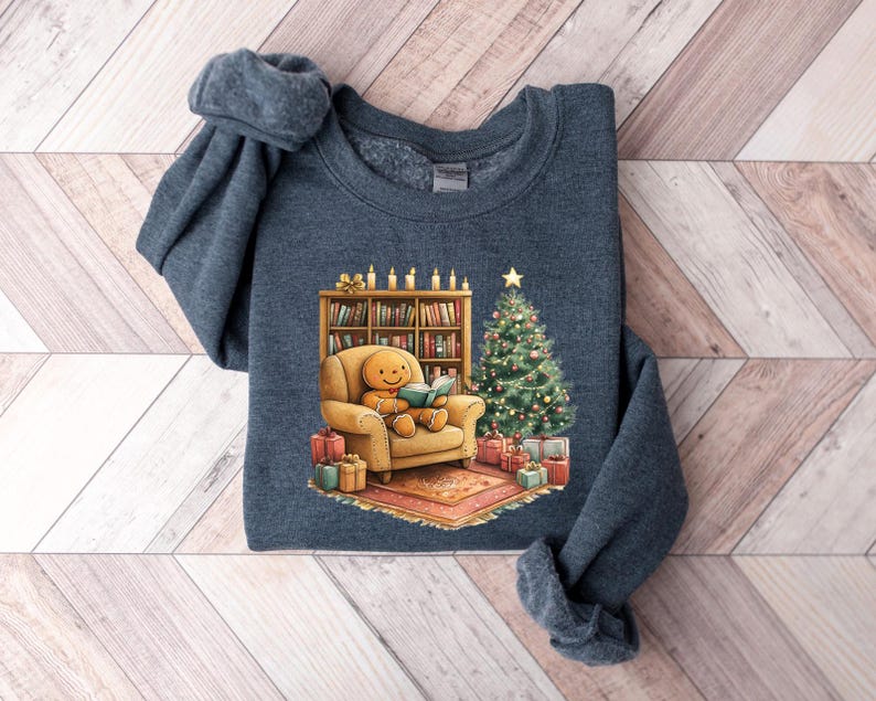 May include: A dark blue sweatshirt with a Christmas-themed design featuring a gingerbread person sitting in a chair reading a book by a fireplace with a Christmas tree and presents.