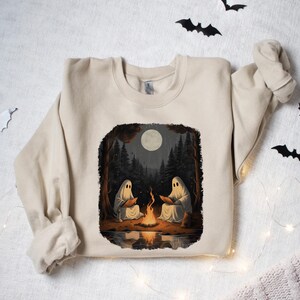 May include: A beige sweatshirt featuring a graphic print of two ghosts sitting by a campfire in a forest. The moon is full and the scene is illuminated by the fire.