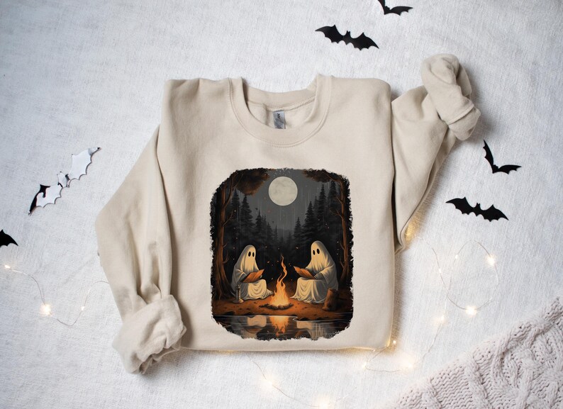 May include: A beige sweatshirt featuring a graphic print of two ghosts sitting by a campfire in a forest. The moon is full and the scene is illuminated by the fire.