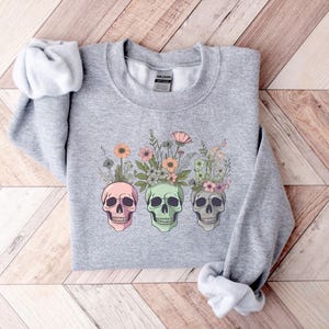 May include: Gray sweatshirt with a floral design featuring three colorful skulls. The flowers are pink, white, and green.