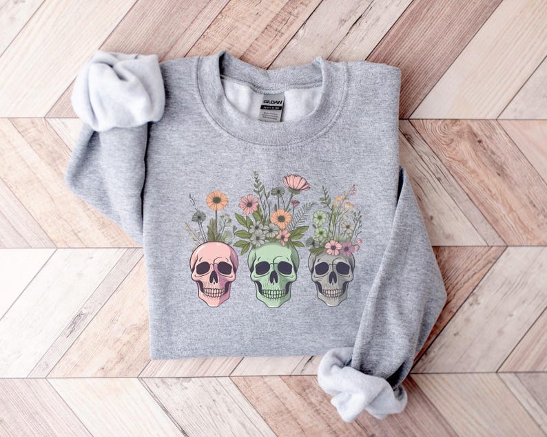 May include: Gray sweatshirt with a floral design featuring three colorful skulls. The flowers are pink, white, and green.