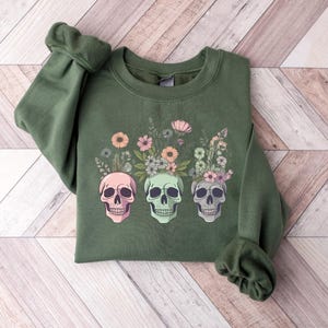 May include: A green sweatshirt with a floral design and three colorful skulls. The skulls are surrounded by flowers and leaves in shades of pink, green, and yellow.