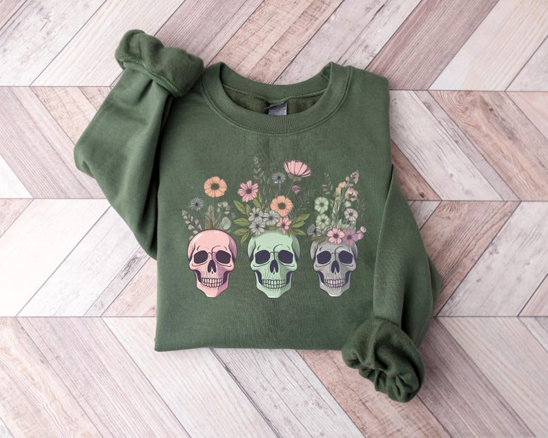 May include: A green sweatshirt with a floral design and three colorful skulls. The skulls are surrounded by flowers and leaves in shades of pink, green, and yellow.