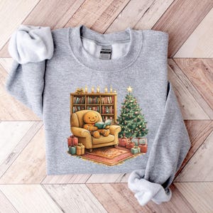 May include: A gray sweatshirt with a Christmas-themed design featuring a gingerbread person sitting in a chair reading a book by a fireplace with a Christmas tree and presents.