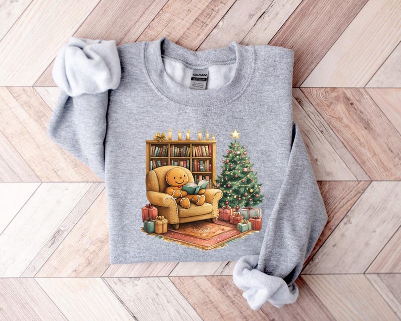 May include: A gray sweatshirt with a Christmas-themed design featuring a gingerbread person sitting in a chair reading a book by a fireplace with a Christmas tree and presents.