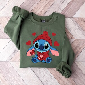Pu&ograve; includere: Una felpa verde oliva con un personaggio dei cartoni animati che indossa un cappello e un maglione rossi, che tiene in mano un lecca-lecca a forma di cuore. Cuori rossi sono sparsi intorno al personaggio. La felpa ha maniche lunghe e girocollo.