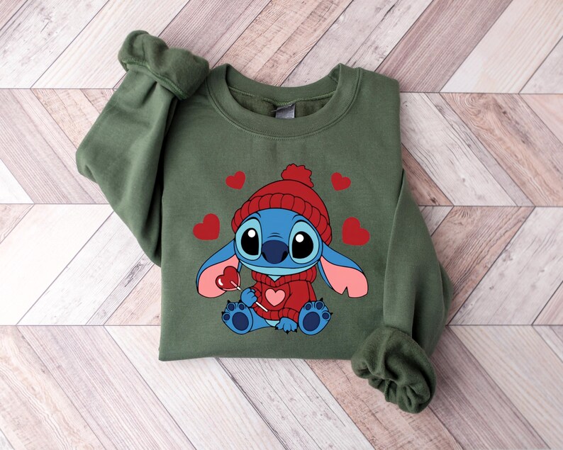 Pu&ograve; includere: Una felpa verde oliva con un personaggio dei cartoni animati che indossa un cappello e un maglione rossi, che tiene in mano un lecca-lecca a forma di cuore. Cuori rossi sono sparsi intorno al personaggio. La felpa ha maniche lunghe e girocollo.