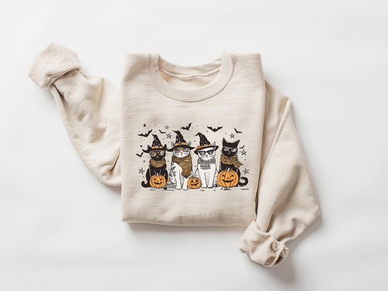 May include: A beige sweatshirt with a graphic print of three cats wearing witch hats and holding pumpkins. The cats are black, white, and gray. The graphic is in a cartoon style.