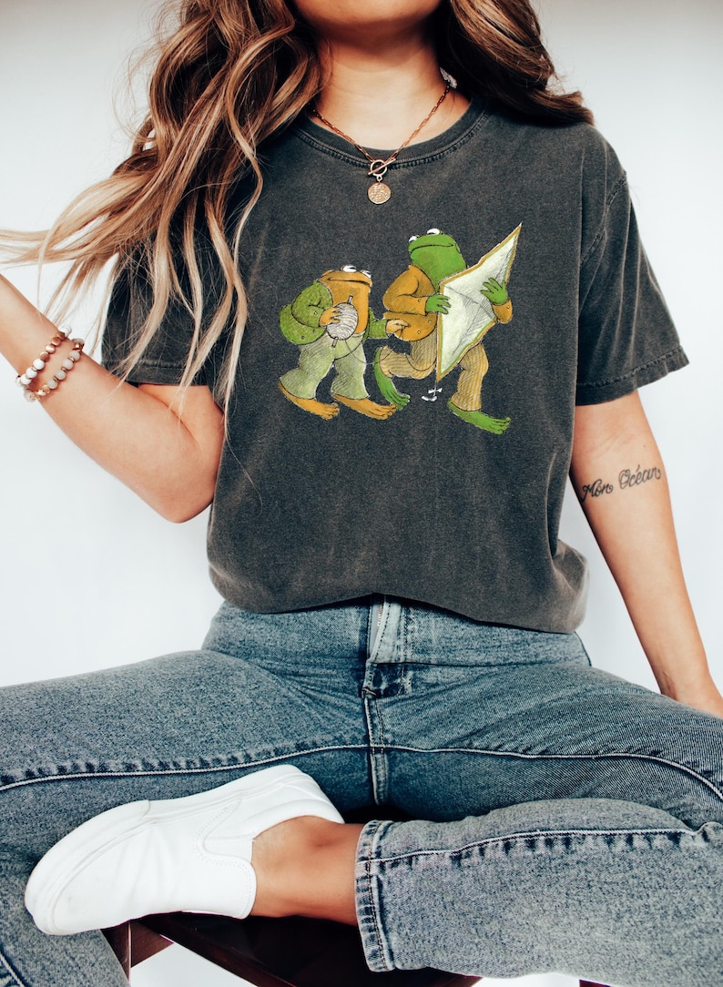 May include: A dark gray t-shirt with a graphic print of two frogs. One frog is holding a ball of yarn and the other is holding a kite. The text "More B&eacute;b&eacute;s" is visible on the right arm.