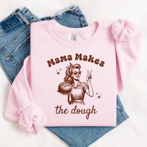 May include: Pink sweatshirt with a brown graphic of a woman holding a loaf of bread and the text "Mama Makes the Dough".