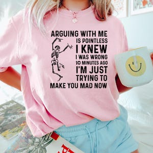 Puede incluir: Sudadera rosa claro con el texto "ARGUING WITH ME IS POINTLESS I KNEW I WAS WRONG 10 MINUTES AGO I'M JUST TRYING TO MAKE YOU MAD NOW" y un gr&aacute;fico de esqueleto. Se sostiene una taza blanca con una carita sonriente amarilla.